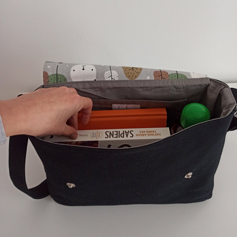 May include: A black canvas messenger bag with a gray lining. The bag is open and contains a book titled "Sapiens", a green water bottle, and a few other items.