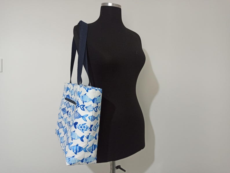 May include: A white tote bag with a blue and white fish pattern. The bag has two navy blue straps.