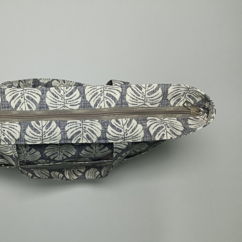May include: A grey and white tote bag with a zip closure and a tropical leaf pattern. The bag has a clear plastic pocket on the front.