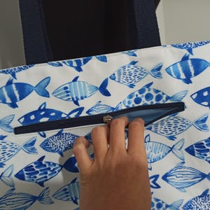 May include: A white canvas tote bag with a blue and white fish pattern. The bag has a zipper closure and navy blue handles.