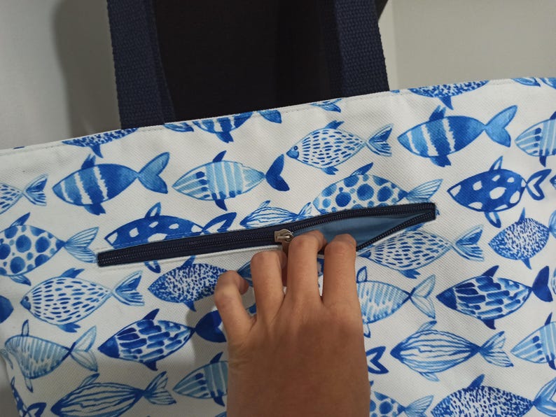 May include: A white canvas tote bag with a blue and white fish pattern. The bag has a zipper closure and navy blue handles.