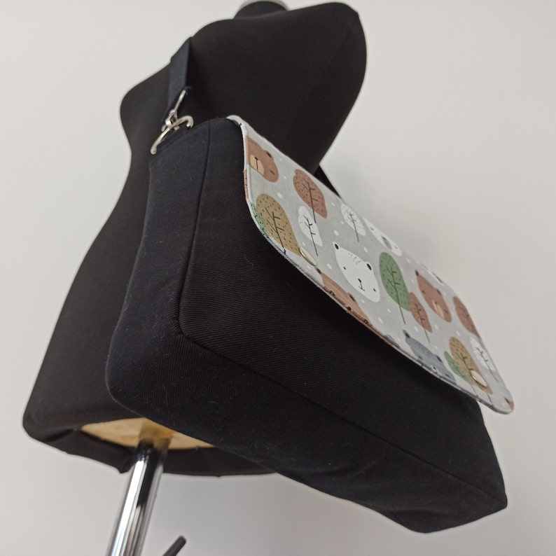 May include: A black canvas messenger bag with a flap closure. The flap is made of a light gray fabric with a pattern of brown bears, trees, and white dots.