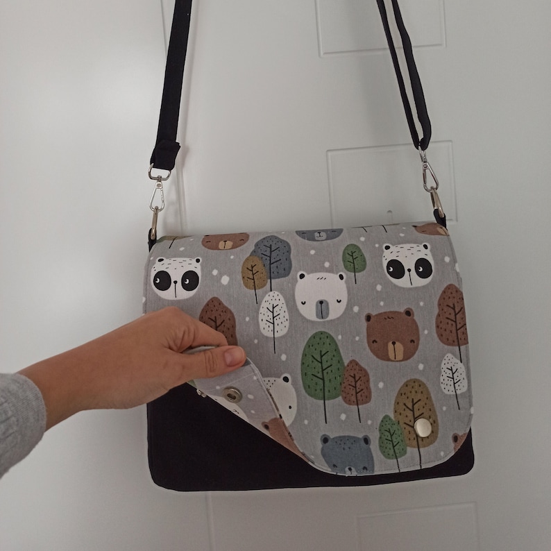 May include: A black and gray shoulder bag with a flap that features a pattern of bears, owls, and trees. The flap is folded down to reveal the black interior of the bag.