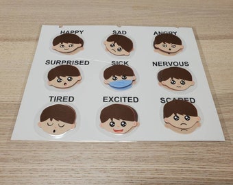Op de afbeelding: Een witte gelamineerde educatieve kaart met negen ronde illustraties van een gezicht dat verschillende emoties toont. Elk gezicht is gelabeld met de bijbehorende tekst: HAPPY, SAD, ANGRY, SURPRISED, SICK, NERVOUS, TIRED, EXCITED en SCARED.