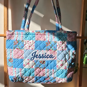 May include: A quilted tote bag featuring a patchwork design of blue, white, and pink floral and gingham patterns. The bag has two matching shoulder straps and the name Jessica is embroidered in blue script on the front center panel.