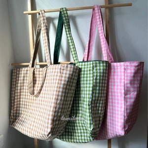 K&ouml;nnte beinhalten: Drei Tragetaschen mit Gingham-Mustern. Die Taschen sind braun, gr&uuml;n und rosa. Die Taschen h&auml;ngen an einem Holzgestell.