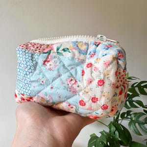 May include: A quilted patchwork pouch with a white zip. The pouch features a variety of floral patterns in shades of red, pink, blue, and white. The fabric is stitched in a diamond pattern.