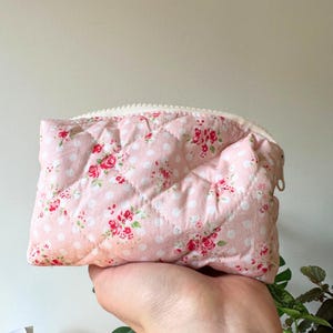 May include: A pink quilted pouch with a floral and polka dot design. It has a white zip and is held in a hand. The floral pattern features red roses and green leaves.