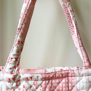 May include: A quilted tote bag with a patchwork design. The bag features a combination of floral, striped, and gingham patterns in shades of pink and white. The handles are made of the same quilted fabric.