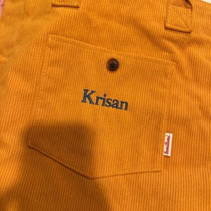 May include: A mustard yellow corduroy tote bag featuring two top handles and a front patch pocket. The pocket is secured with a single brown button and displays the embroidered blue text Krisan, with a small white tag labeled tomitomi.