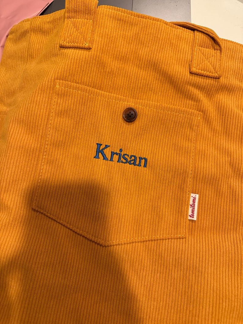 May include: A mustard yellow corduroy tote bag featuring two top handles and a front patch pocket. The pocket is secured with a single brown button and displays the embroidered blue text Krisan, with a small white tag labeled tomitomi.