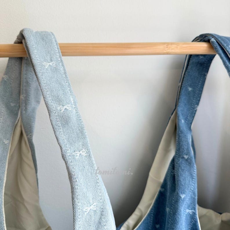 May include: Close-up of two denim tote bag straps hanging from a wooden rod. The straps are light blue denim with a white bow pattern. The interior of the bag is a light beige color. The bags are hanging against a white wall.