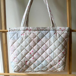 May include: Quilted tote bag with floral and checkered patchwork design in pastel pink, blue, and white. The bag features quilted diamond pattern and matching handles. A charming and practical accessory.