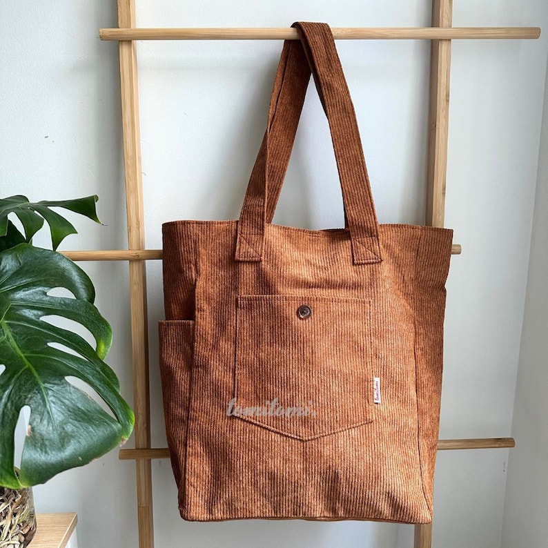 May include: A rust-colored corduroy tote bag featuring two long shoulder straps, a large front pocket secured with a single brown button, and a small white rectangular brand label on the side. The fabric has a distinct vertical ribbed texture.