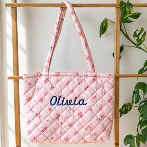 Op de afbeelding: Een gewatteerde tote bag van lichtroze stof met een klein rood bloemenpatroon. De tas heeft twee schouderbanden en de naam Olivia is in donkerblauw garen geborduurd in het midden van het voorpaneel van de gewatteerde tas.