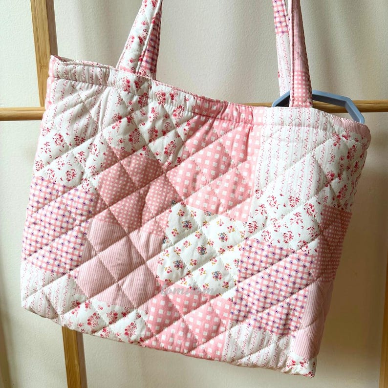 May include: A quilted tote bag with a patchwork design. The bag features a variety of pink and white fabrics, including floral prints, polka dots, and gingham patterns. The bag has two handles and is made of a soft, padded material.