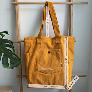 May include: A mustard yellow corduroy tote bag with two shoulder straps, a front pocket with a button, and side pockets. The bag measures 16.1 inches wide, 15.4 inches high, 3.9 inches deep, with a 10.2-inch strap drop. The text 'tomitomi' is visible.