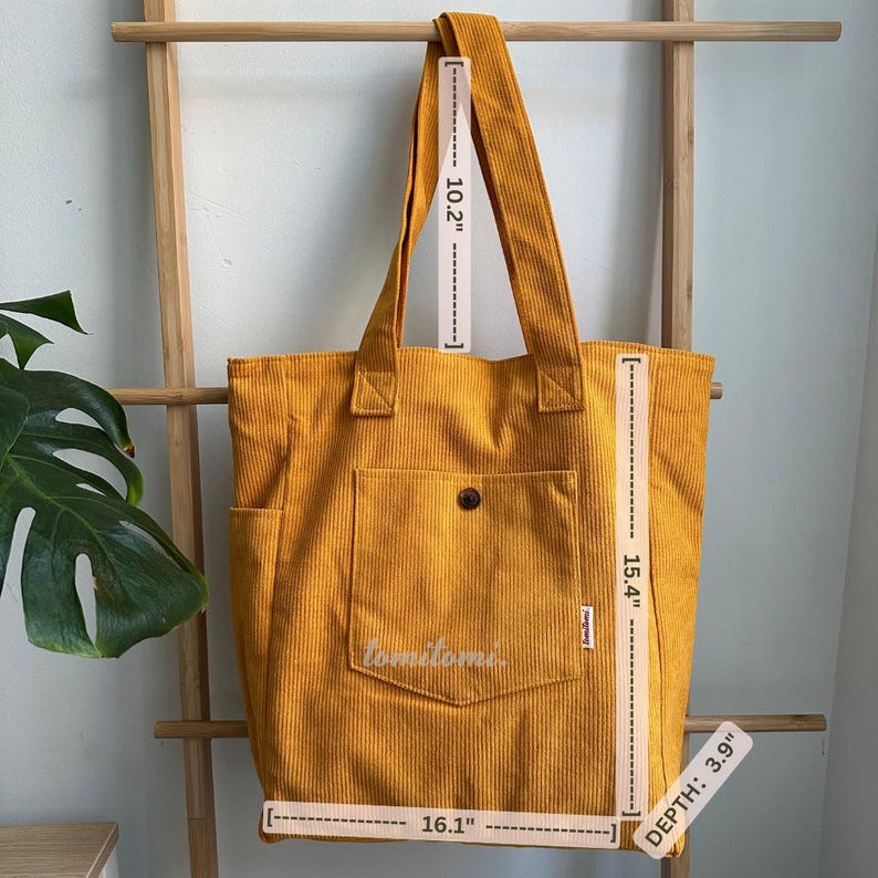 May include: A mustard yellow corduroy tote bag with two shoulder straps, a front pocket with a button, and side pockets. The bag measures 16.1 inches wide, 15.4 inches high, 3.9 inches deep, with a 10.2-inch strap drop. The text 'tomitomi' is visible.