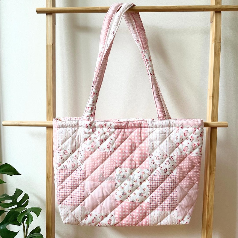 May include: A quilted tote bag with a patchwork design in shades of pink and white. The bag features a variety of floral and geometric patterns, with sturdy handles for carrying. The bag is displayed on a wooden rack.