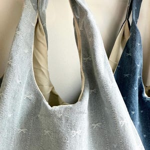 May include: Two denim tote bags with a light blue wash and a bow pattern. The bags have a cream-colored lining and long straps. One bag is a lighter shade of blue than the other.