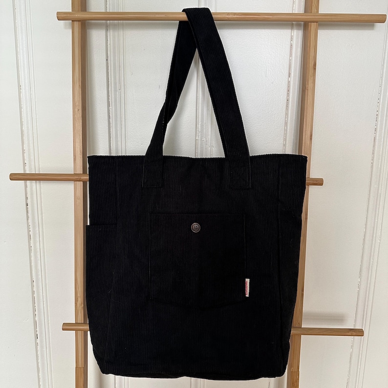 May include: A black corduroy tote bag featuring two shoulder straps, a large front pocket secured with a single brown button, and a small rectangular white fabric tag with red text attached to the side of the front pocket.