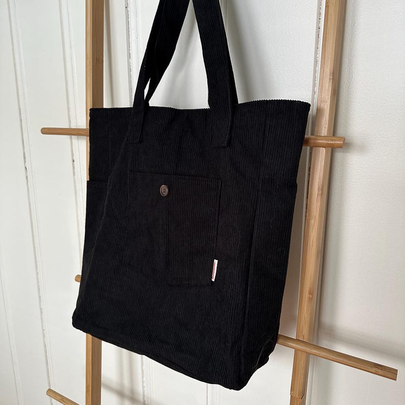 May include: A black corduroy tote bag featuring a vertical ribbed texture, two shoulder straps, and a central exterior pocket secured with a single brown button. A small white rectangular fabric tag is attached to the side of the front pocket.