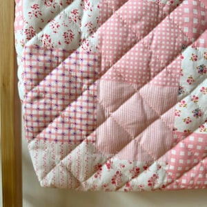 May include: A quilted patchwork item featuring a variety of pink and white floral, gingham, and dotted patterns. The diagonal design is stitched with a diamond pattern. The item is likely a decorative piece or part of a larger textile project.