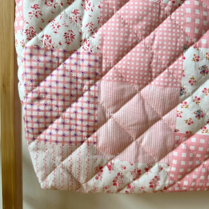 May include: A quilted patchwork item featuring a variety of pink and white floral, gingham, and dotted patterns. The diagonal design is stitched with a diamond pattern. The item is likely a decorative piece or part of a larger textile project.