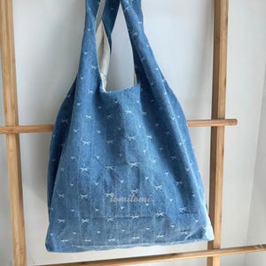 May include: A blue denim tote bag with white bow patterns. The bag has a large capacity and long straps for shoulder or hand carry. The bag has the text "tomitomi" on the front.