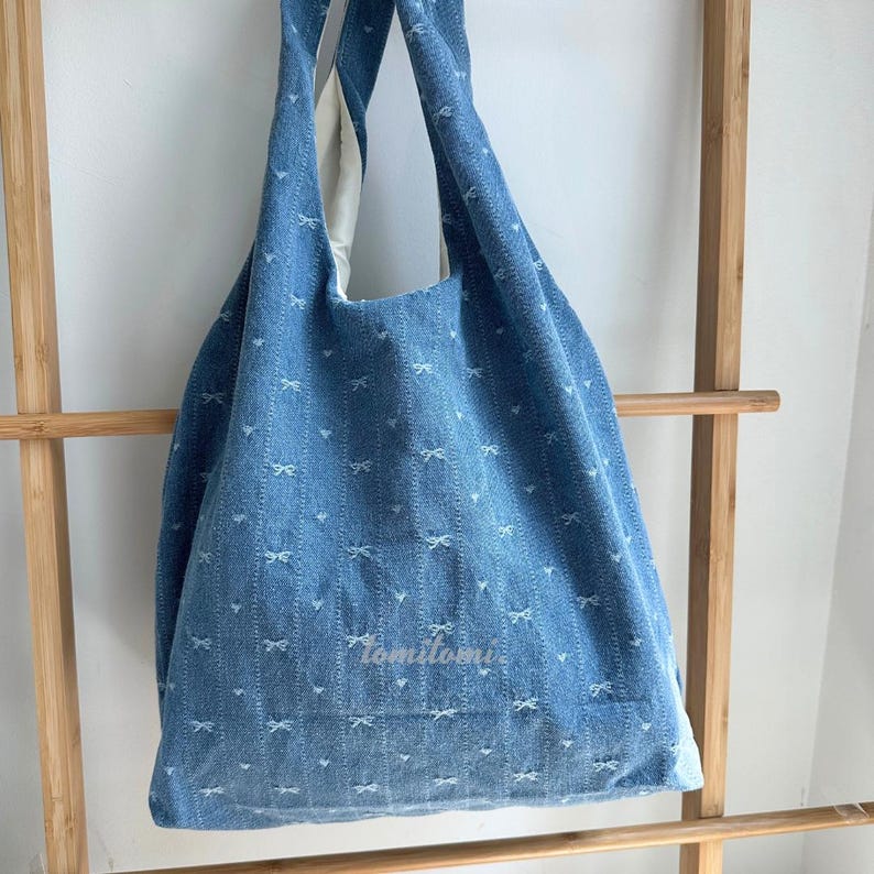 May include: A blue denim tote bag with white bow patterns. The bag has a large capacity and long straps for shoulder or hand carry. The bag has the text "tomitomi" on the front.