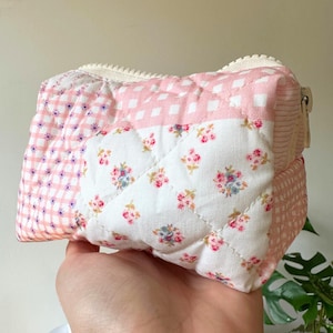 Op de afbeelding: Een gewatteerde make-uptas met een patchworkontwerp. De tas heeft delen van roze ruitjes, bloemenprint en een roze en wit ruitpatroon. Het heeft een crèmekleurige ritssluiting en wordt in een hand gehouden.