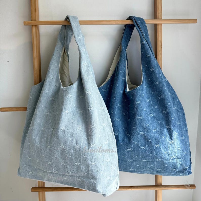 May include: Two denim tote bags, one light blue and one dark blue, hang on a wooden rack. Both bags have a repeating bow pattern and a wide shoulder strap. The light blue bag has the word "itomi" printed on it.