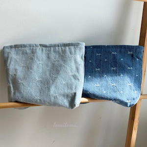 May include: Two denim fabric pouches, one light blue and one dark blue, each with a repeating bow pattern. The pouches are displayed on a wooden shelf against a white wall. The light blue pouch is on the left, and the dark blue pouch is on the right.