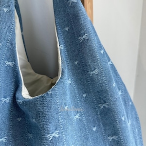 May include: Close-up of a light blue denim tote bag with a white interior. The bag features a repeating pattern of small white bows and hearts. The bag is made of a sturdy denim material.