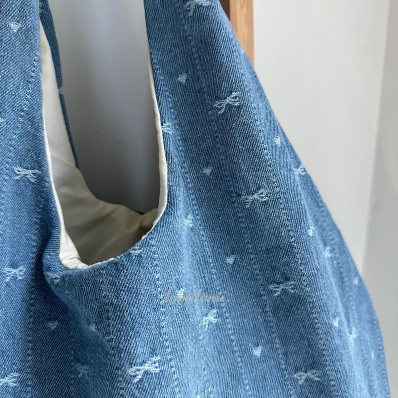 May include: Close-up of a light blue denim tote bag with a white interior. The bag features a repeating pattern of small white bows and hearts. The bag is made of a sturdy denim material.