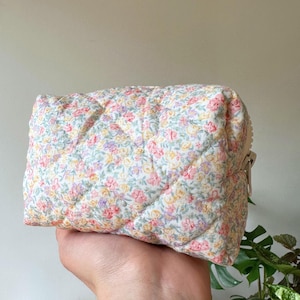 May include: A quilted cosmetic bag with a floral print. The bag features a pattern of pink, yellow, and purple flowers on a white background. It has a rectangular shape and a silver zip closure. The bag is made of fabric.