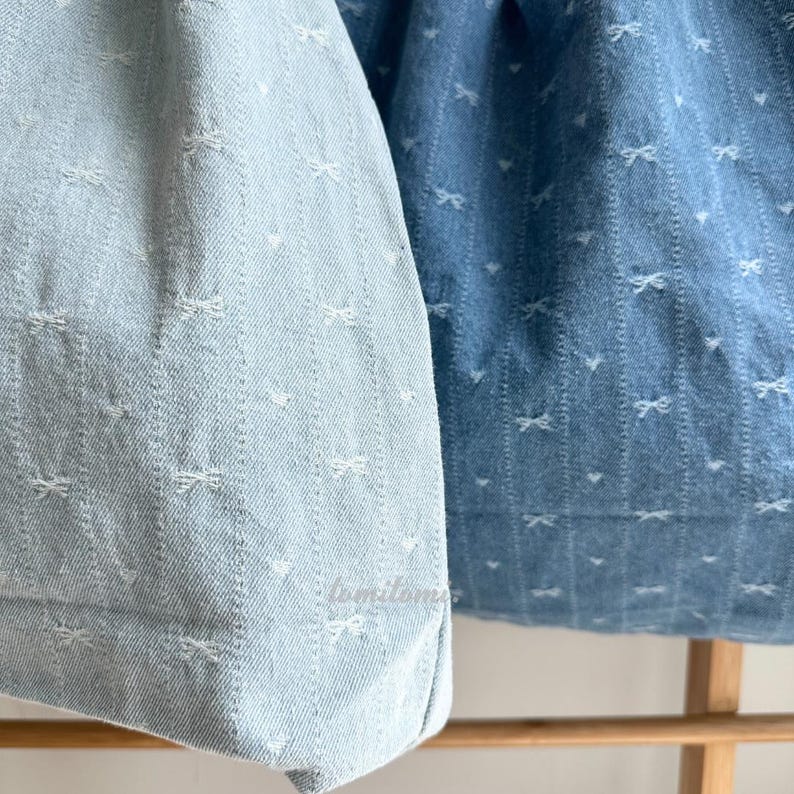 May include: Close-up of two denim fabric pieces, one light blue and one medium blue. Both feature a repeating pattern of small white bows and vertical lines, creating a textured effect. The fabric appears to be soft and lightweight.