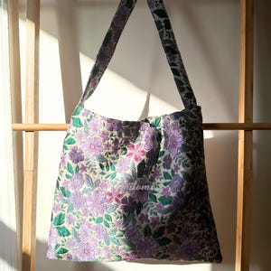 May include: A floral tote bag with a quilted design. The bag features a pattern of purple, pink, and green flowers on a cream background. The shoulder strap and bag lining have a coordinating floral pattern.