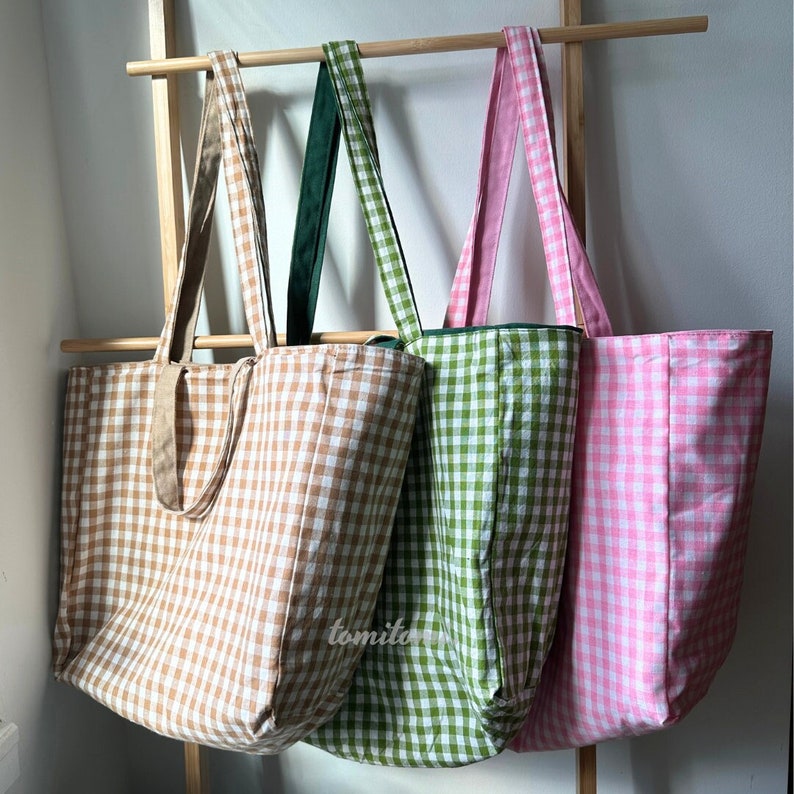 K&ouml;nnte beinhalten: Drei Tragetaschen mit Gingham-Mustern. Die Taschen sind braun, gr&uuml;n und rosa. Die Taschen h&auml;ngen an einem Holzgestell.