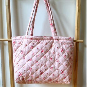 May include: A pink quilted tote bag with a floral pattern. The bag features a repeating pattern of small red flowers and green leaves on a light pink background. The bag has two long handles and a diamond-shaped quilted design.