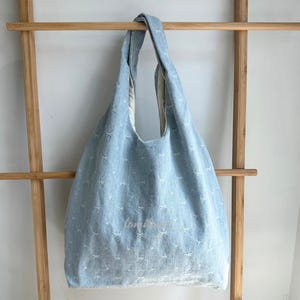 May include: Light blue denim tote bag with white embroidered star patterns. The bag has a wide opening and long handles. The text "tonight" is embroidered on the front. The bag is hanging on a wooden rack.