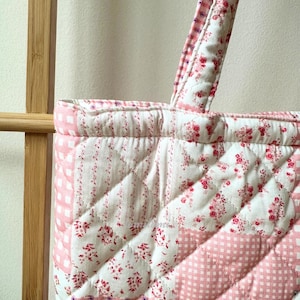 May include: Close-up of a quilted tote bag with a floral and gingham pattern in pink and white. The bag features a quilted handle and a diamond pattern. The fabric has a vintage, cottagecore aesthetic.