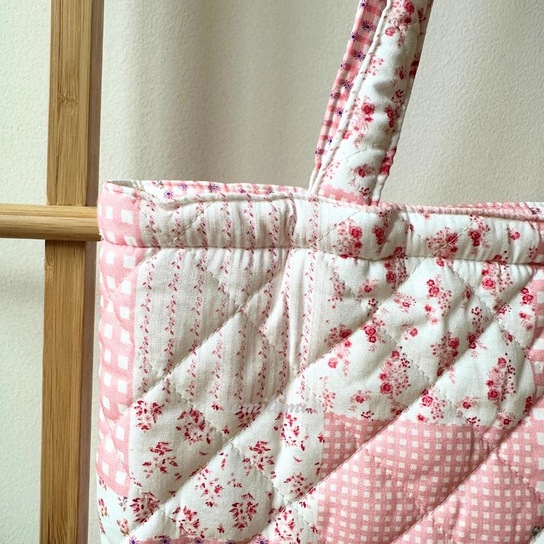 May include: Close-up of a quilted tote bag with a floral and gingham pattern in pink and white. The bag features a quilted handle and a diamond pattern. The fabric has a vintage, cottagecore aesthetic.