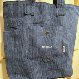 May include: A blue corduroy tote bag featuring two top handles, a large front pocket with a single brown button closure, and the text HARTLEY embroidered in white. A small red and white tag with the text tomitomi is attached to the pocket.