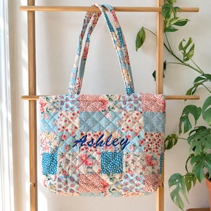 Op de afbeelding: Een gewatteerde tote bag met een patchworkontwerp van lichtblauwe, roze en witte bloemen- en stippenpatronen. De tas heeft twee bijpassende gewatteerde schouderbanden en de naam Ashley is in donkerblauw cursief schrift op het midden geborduurd.