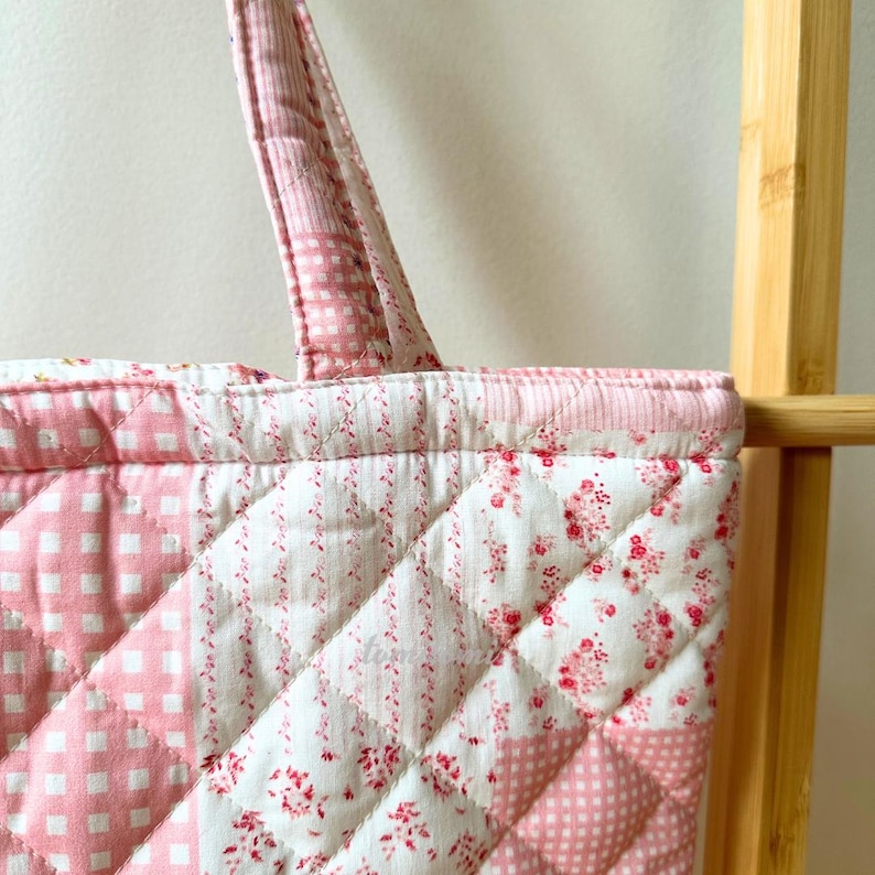 May include: A quilted tote bag with a patchwork design. The bag features various pink and white patterns, including gingham, floral, and polka dots. The bag has a quilted texture and a sturdy handle for carrying. The overall aesthetic is vintage and feminine.