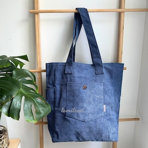 May include: A blue corduroy tote bag featuring a vertical ribbed texture, two shoulder straps, and a large front pocket secured with a single brown button. The word tomitomi is printed in white on the front pocket, and a small tag is attached.