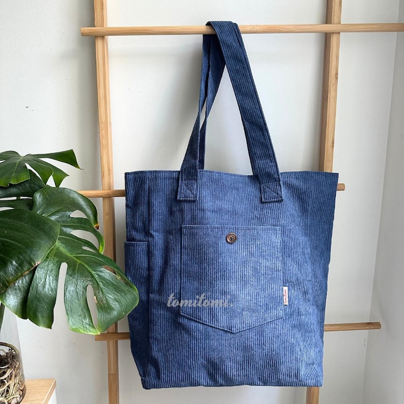 May include: A blue corduroy tote bag featuring a vertical ribbed texture, two shoulder straps, and a large front pocket secured with a single brown button. The word tomitomi is printed in white on the front pocket, and a small tag is attached.