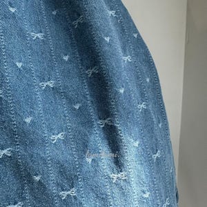 May include: Close-up of blue denim fabric with a pattern of small white bows and vertical lines. The fabric has a slightly distressed appearance, with some areas showing lighter shades of blue.