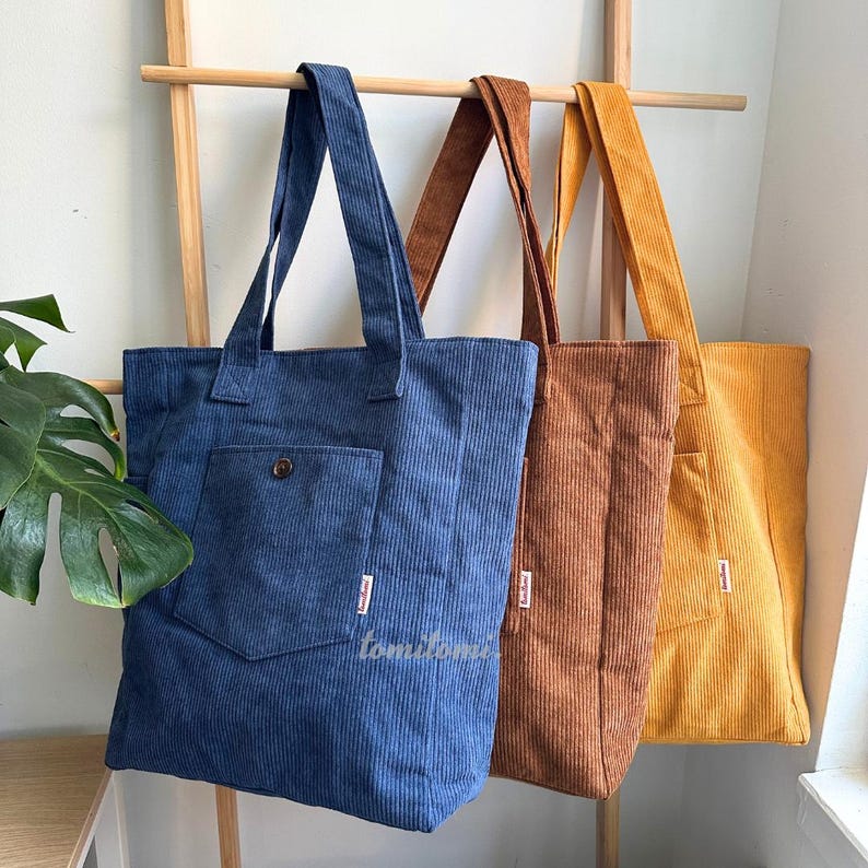 May include: Three corduroy tote bags in blue, brown, and yellow hanging on a wooden rack. Each bag features a large front pocket with a single button closure and a small white rectangular label on the side. The fabric has a vertical ribbed texture.